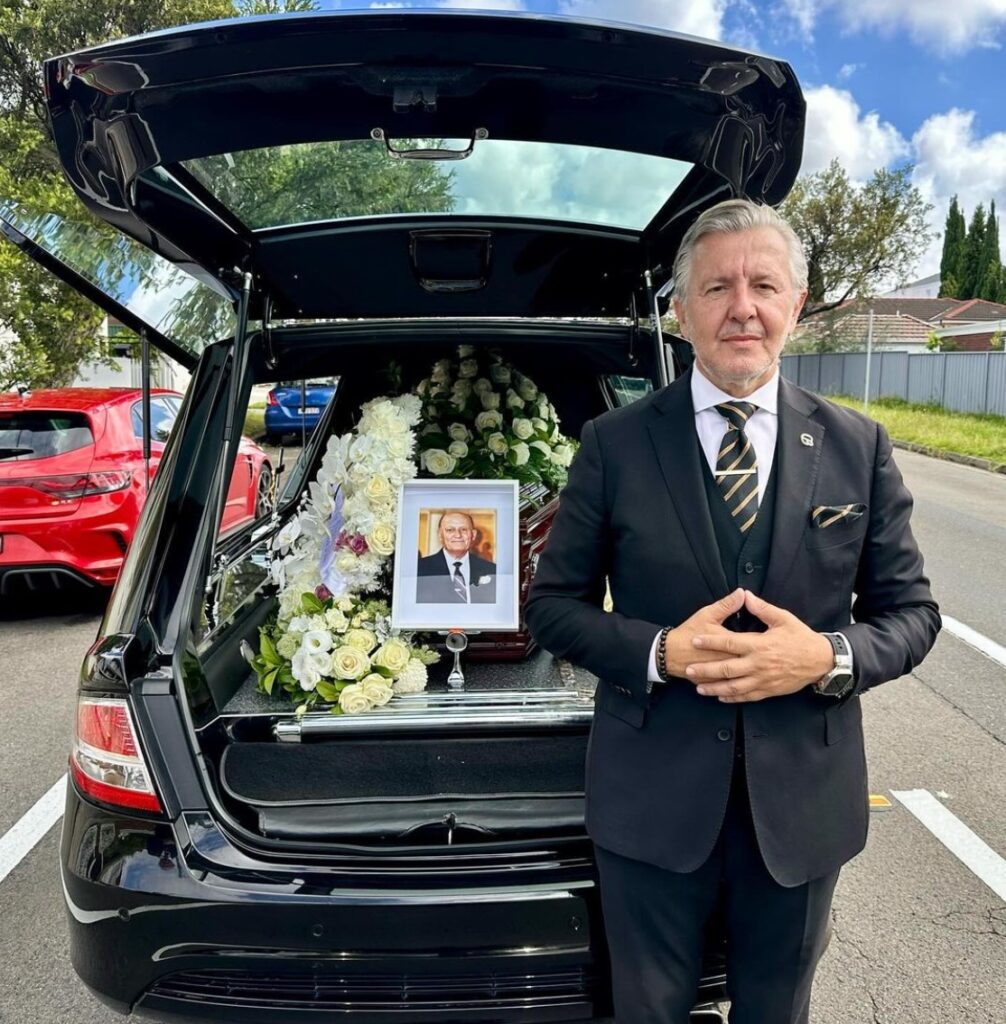 Jim Georges, Director at Divine Family Funerals, standing behind an open hearse with a casket inside