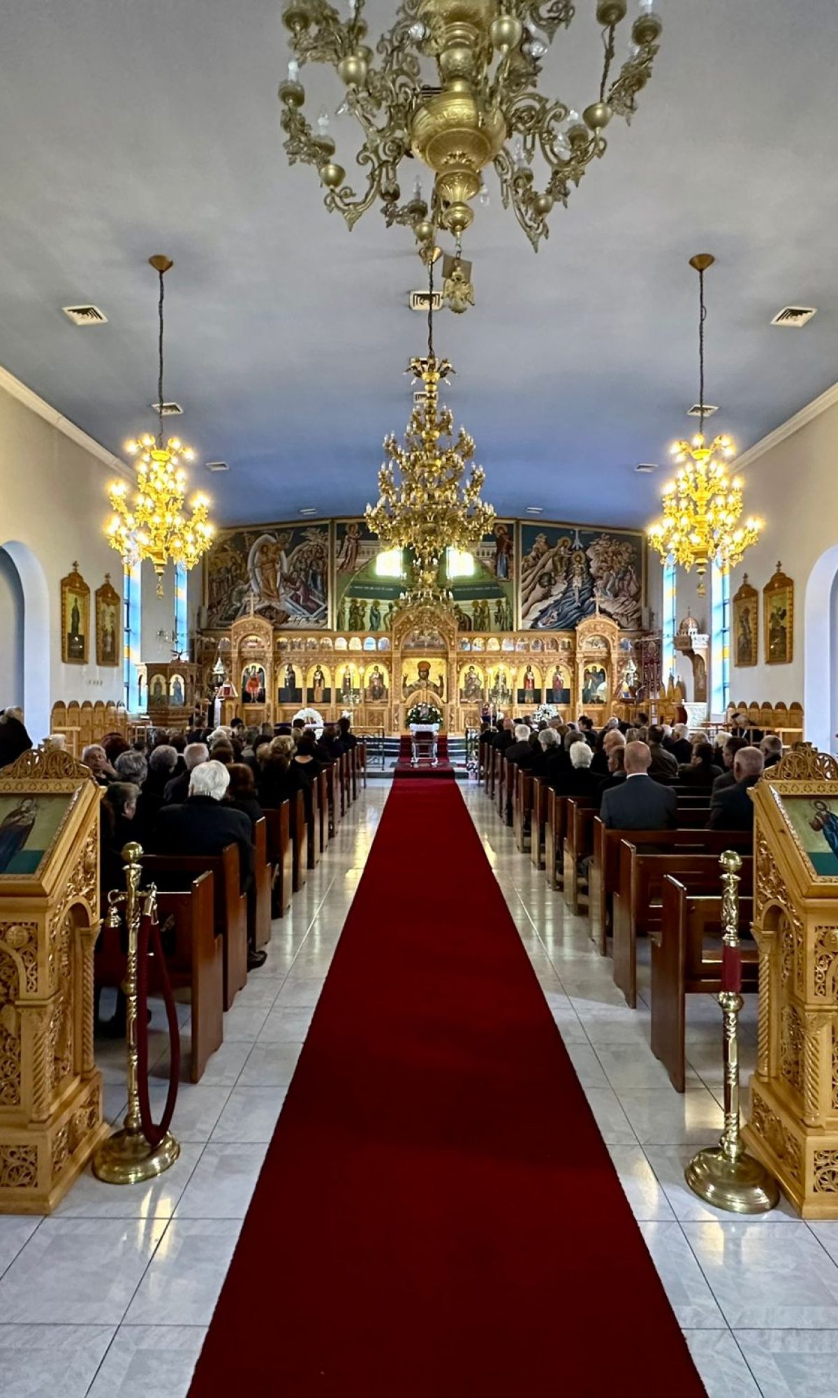 Rear view inside a Greek Orthodox church during a funeral service, showing the congregation and the coffin placed at the front.